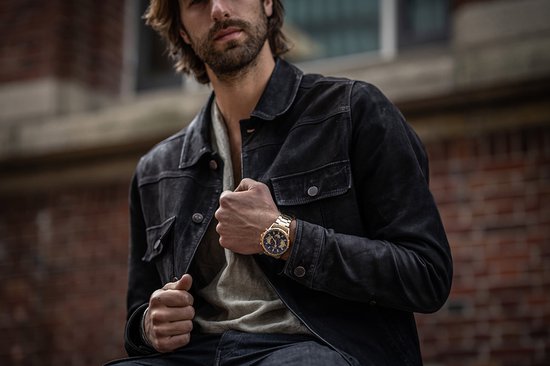 Man met een donker jack en een gouden horloge met blauwe wijzerplaat om zijn pols.