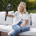 Vrouw met blond haar, witte blouse en spijkerbroek, zittend op een bank met een selfiestick in haar hand.