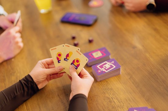 Een persoon houdt kaarten van "Music Match" vast met tekst zoals "VOCALS" en "LAND" in handen. Op tafel liggen paarse stapels kaarten.