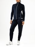 Model in een volledig donkerblauw trainingspak. Het jack heeft een witte bies en de broek heeft een witte bies langs de pijpen. Merk: Sjeng Sports.