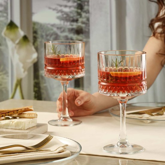 Twee hoge cocktailglazen met een brede voet, verticale ribbels en een diamantpatroon, elk gevuld met een rode drank, sinaasappelschijfjes en rozemarijn.