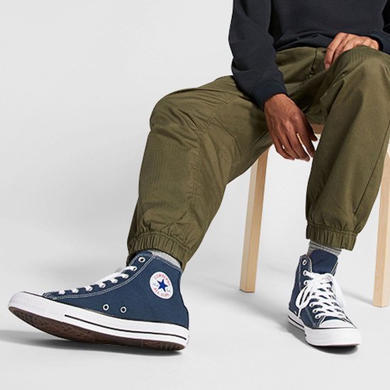 Man zittend op een stoel met donkerblauwe Converse hoge sneakers aan, met groene broek.