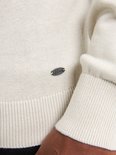 Close-up van een beige trui met ronde hals en klein metalen logo.