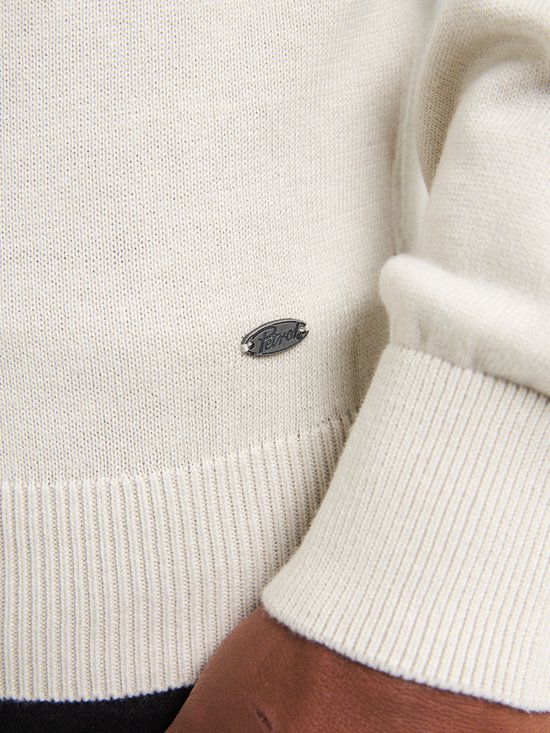Close-up van een beige trui met ronde hals en klein metalen logo.