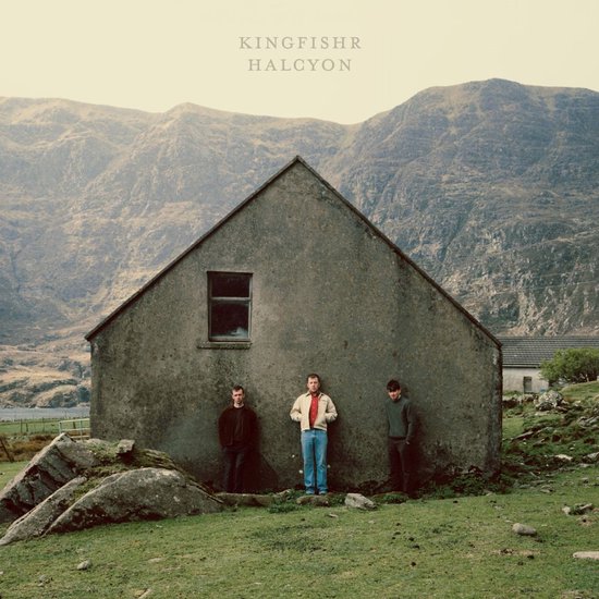 Drie mannen staan voor een stenen huis bij bergen. Albumhoes Kingfishr Halcyon met ingetogen kleding.