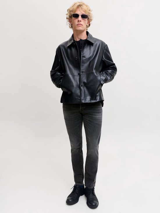Model met blond haar en zonnebril draagt een grijze slim fit jeans en een zwarte leren jas.