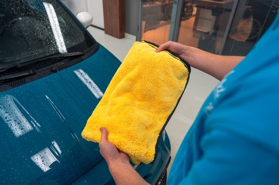 Een teal blauwe auto met waterdruppels op de motorkap, terwijl een persoon een felgeleende microvezel droogdoek vasthoudt.