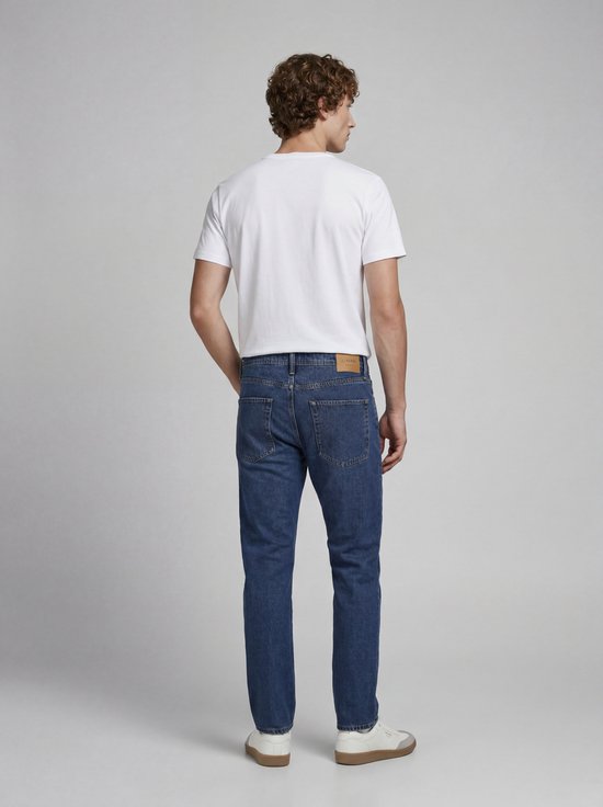 Jongeman in een witte T-shirt met 'JJ REBEL' opdruk en een blauwe spijkerbroek, achterkant.