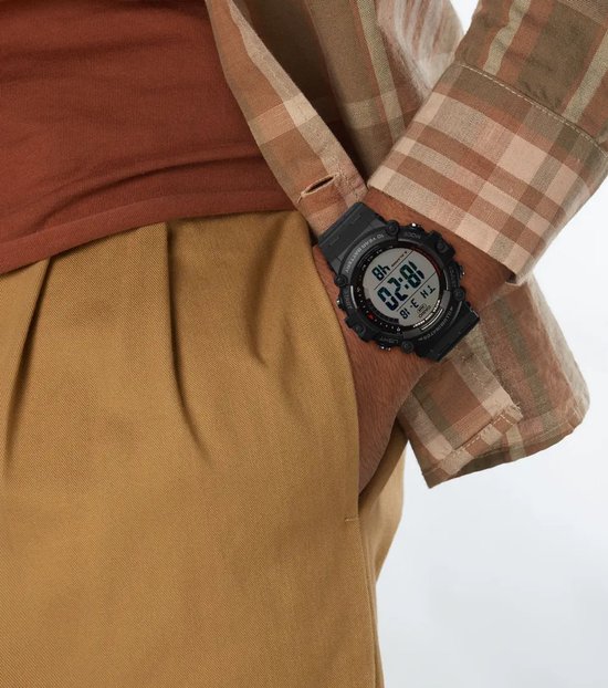 Zwart Casio digitaal horloge met ronde kast om een pols, gedragen bij een geruit overhemd en bruine broek.