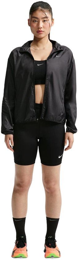Model van voren in een zwarte Nike hardloopjas, zwarte sportbeha, korte broek en kleurrijke schoenen.