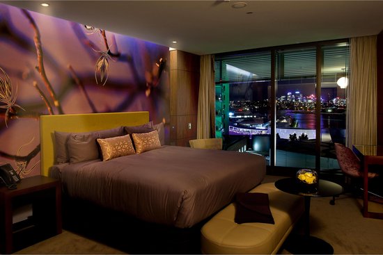 Luxe slaapkamer met groot bed, paars beddengoed, wanddecoratie met takken en uitzicht op de stad.