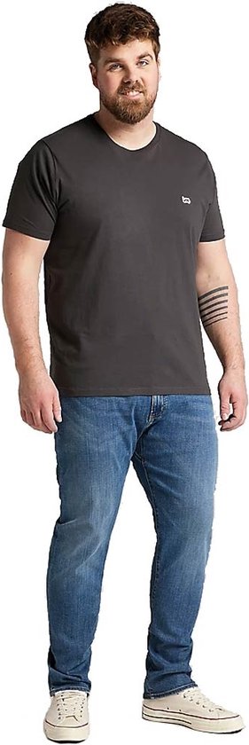 Grote man met een donkergrijs T-shirt en een lichtblauwe jeans, staand met handen in de zakken.