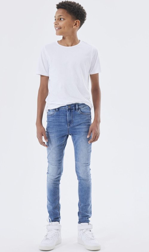 Jongensmodel in witte sneakers, een witte t-shirt en een skinny spijkerbroek van medium blue denim.