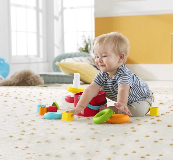 Baby speelt met Fisher-Price stapelpiramide en blokjes op een kleed. Naast de piramide liggen losse gekleurde ringen en blokjes.