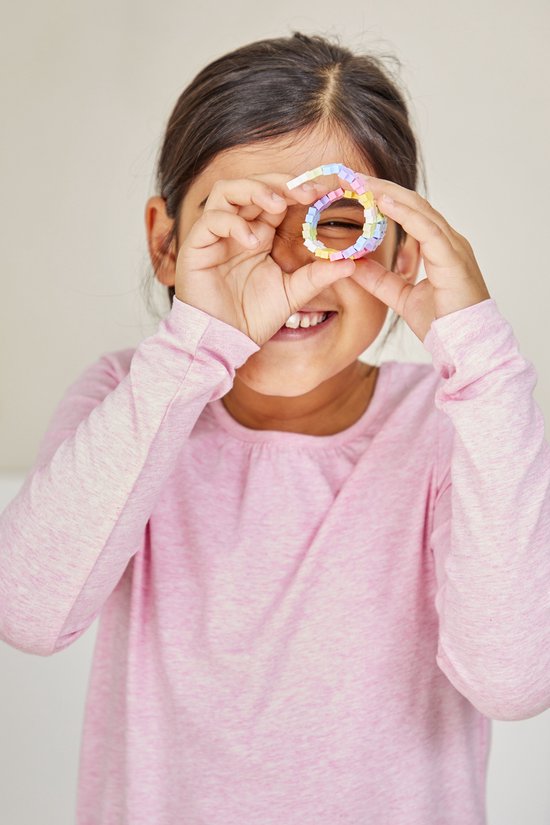 Een jong meisje met een roze trui houdt een regenboogkleurig Plus-Plus bouwwerk in een cirkel voor haar oog.
