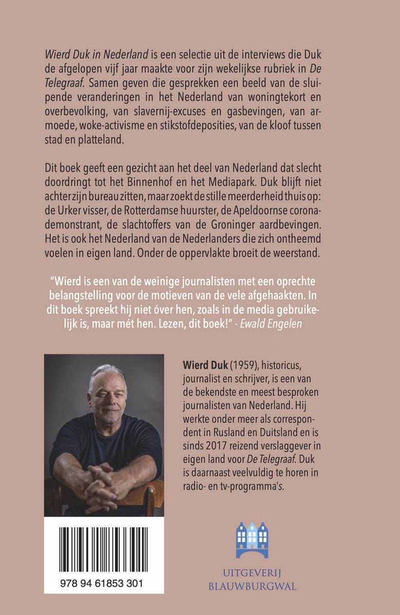 Foto van een man met grijs haar die op een stoel zit met een boek in zijn handen. Hij draagt een donkerblauwe jas en een wit shirt.