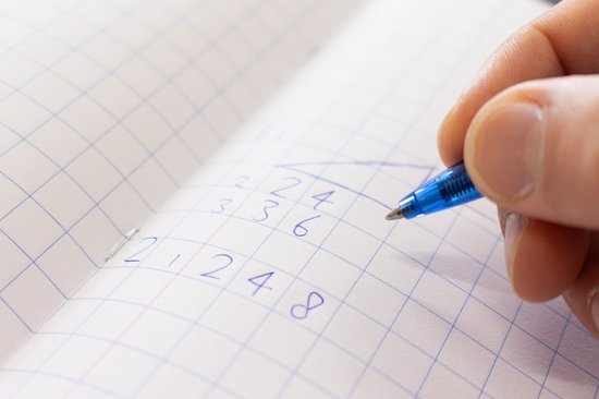 Hand schrijft met blauwe pen in een open geruit schrift. Getallen zoals 2 4, 3 6, 2 4 8 zijn zichtbaar.