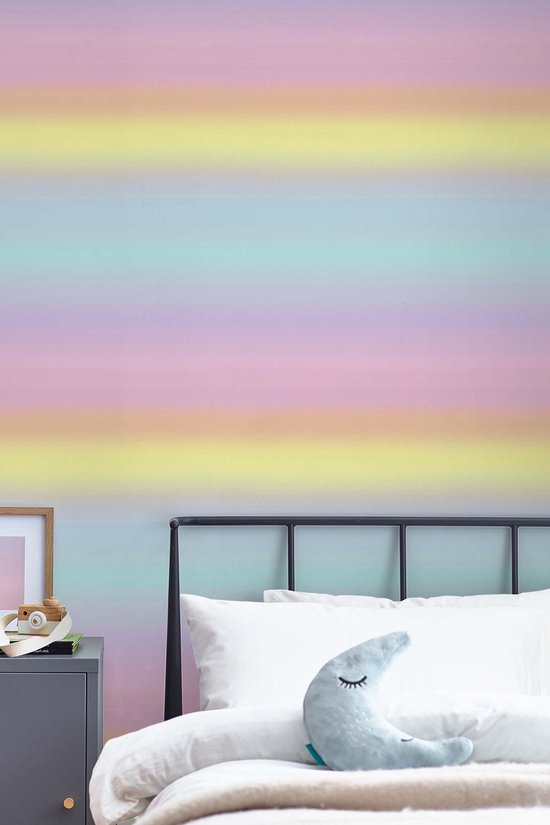 Pastelkleurig regenboogbehang aangebracht op een muur boven een bed met witte kussens en een maan-kussen.