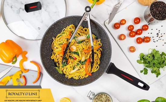 Poêle wok grise mouchetée de noir, remplie de nouilles et de légumes, tenue par une pince. Des poivrons, des tomates cerises et des herbes sont à côté de la poêle.