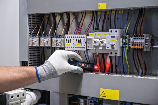 Hand met handschoen houdt een blauwe en zwarte Bosch spanningstester bij een rij automaten.