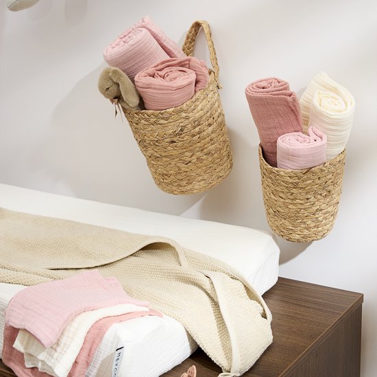 Babykamer met opgerolde zachtroze en crèmekleurige hydrofiele doeken in manden en op een commode.