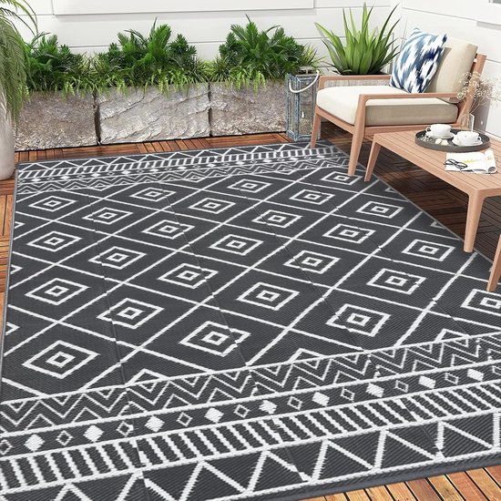Tapis d'extérieur gris foncé avec motif losange blanc et bordures géométriques. Placé sur une terrasse en bois avec des chaises et une table.
