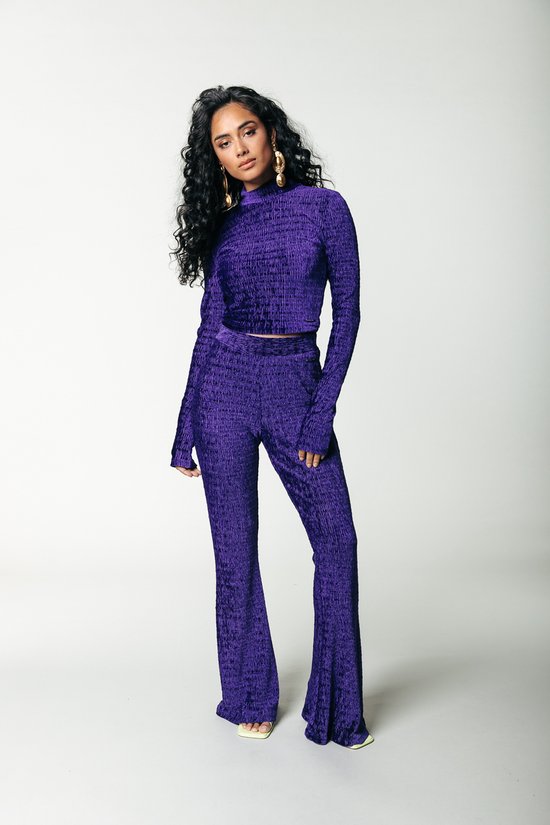 Femme aux cheveux bouclés portant un haut court col roulé en velours violet à manches longues et un pantalon évasé, avec de grandes boucles d'oreilles dorées.