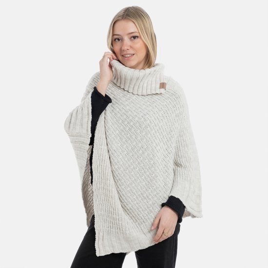 Lichtbeige gebreide poncho met hoge sjaalkraag, gedragen door een blonde vrouw met zwarte coltrui.