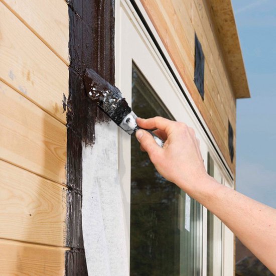 Hand strijkt donkerbruine, rubberachtige coating op houten gevelbekleding naast een raam.