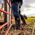 Zwarte Muckmaster laarzen gedragen door persoon, leunend op hek, met quad op achtergrond.