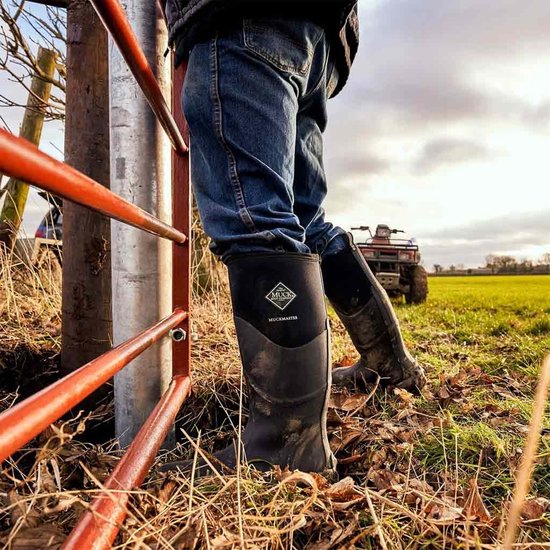 Zwarte Muckmaster laarzen gedragen door persoon, leunend op hek, met quad op achtergrond.