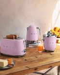 Roze SMEG broodrooster, waterkoker en citruspers op een houten tafel met sinaasappels.