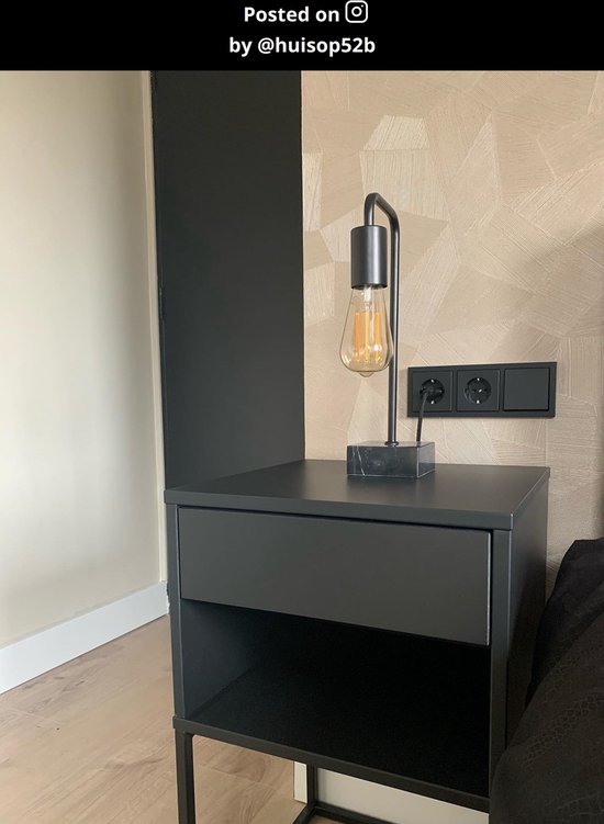 Zwarte tafellamp met gloeilamp op een zwart nachtkastje naast een bed.