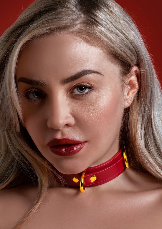 Close-up van een model met blond haar dat een rode halsband met goudkleurige D-ringen draagt.