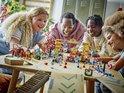 Een diverse groep lachende mensen bouwt aan Lego-raceauto's en figuren rondom een start/finishboog met "Mario Kart" erop.
