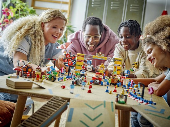 Een diverse groep lachende mensen bouwt aan Lego-raceauto's en figuren rondom een start/finishboog met "Mario Kart" erop.