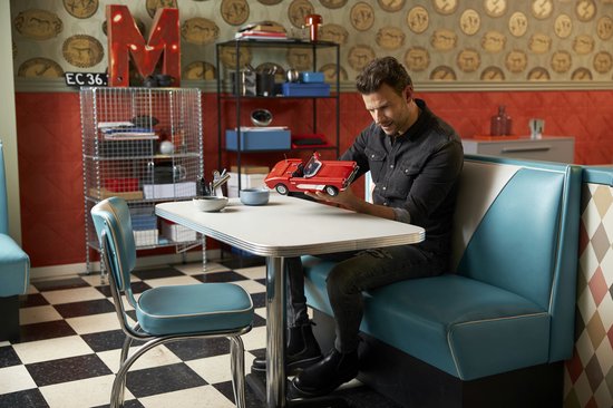 Een man zit aan een tafel in een retro diner, met een rode LEGO Chevrolet Corvette cabriolet in zijn handen. De auto heeft een wit interieur.