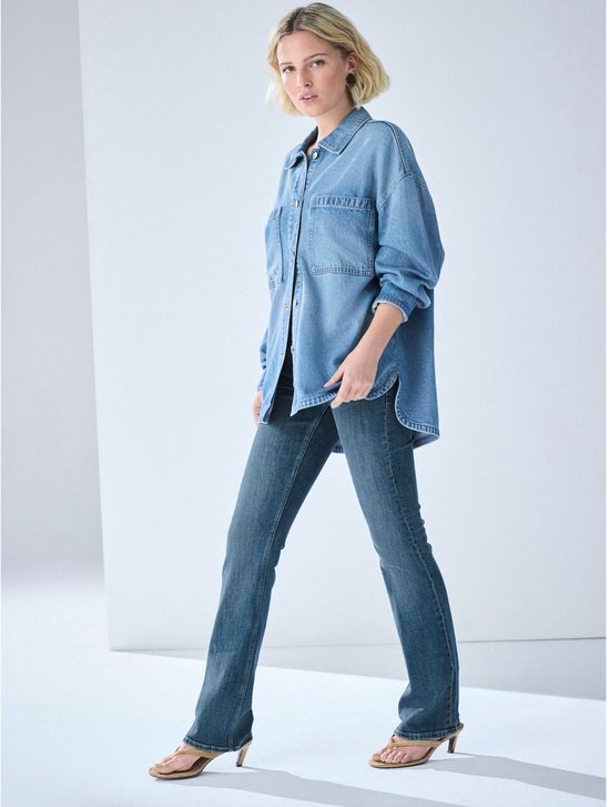 Model staat en draagt een losse denim blouse en een bootcut spijkerbroek met middelhoge taille.