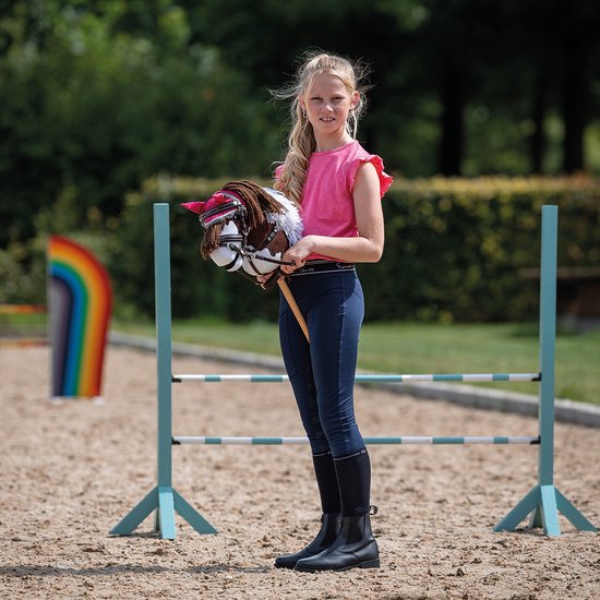Meisje met blonde staartjes houdt bruin-witte hobbyhorse met roze strik vast voor regenbooghindernis.
