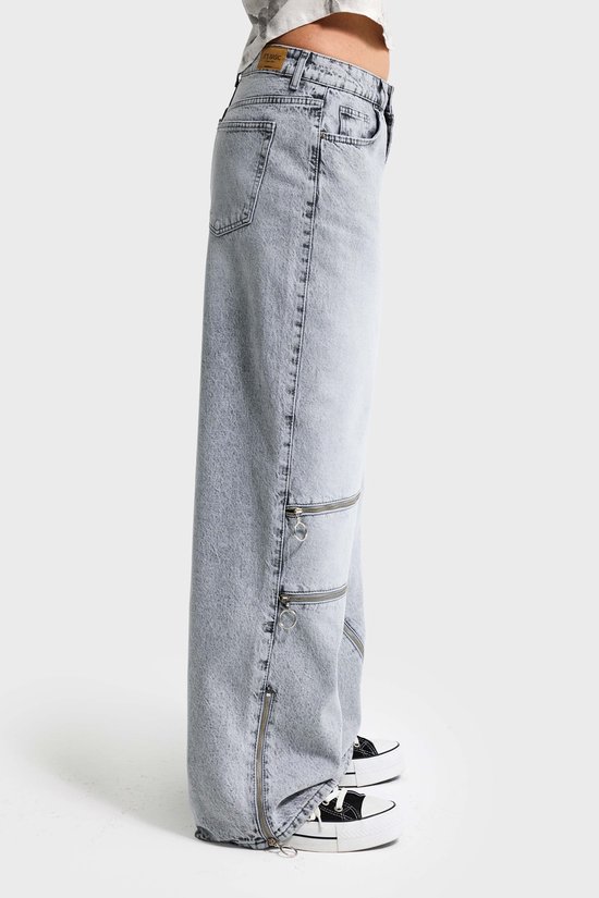 Zijaanzicht van lichtgrijze baggy jeans met ritsdetails op de pijpen.