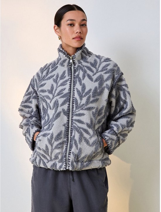 Model draagt een grijs borg-jack met een bloemenprint, ritssluiting en opstaande kraag, gecombineerd met een grijze broek.