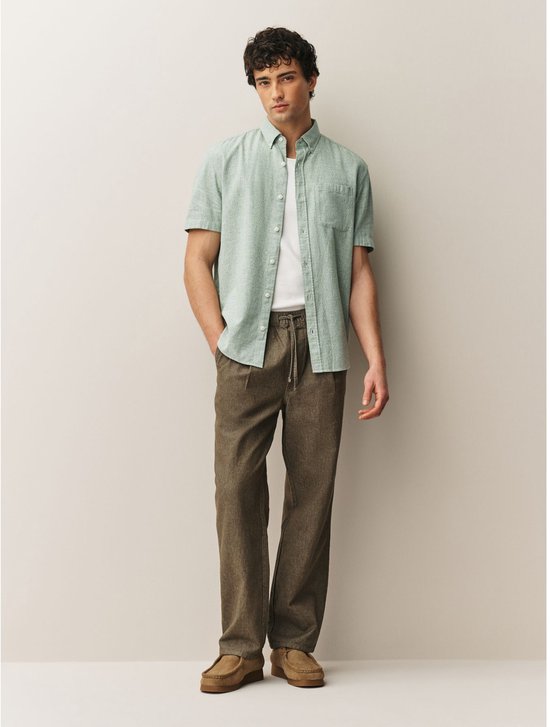 Man met korte mouwen lichtgroen geblokt overhemd, wit T-shirt, bruine broek.