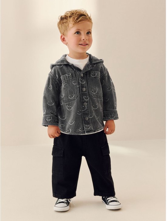 Jongen in grijs denim overshirt met capuchon, smiley-print, zwarte broek en sneakers.