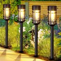 Vier zwarte solar tuinlampen met vlam-effect LED verlichting in een tuin.