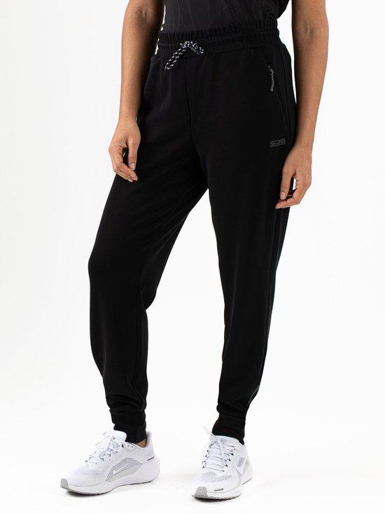 Model in zwarte trainingsbroek met trekkoord, ritszak en logo, gedragen met witte sneakers.