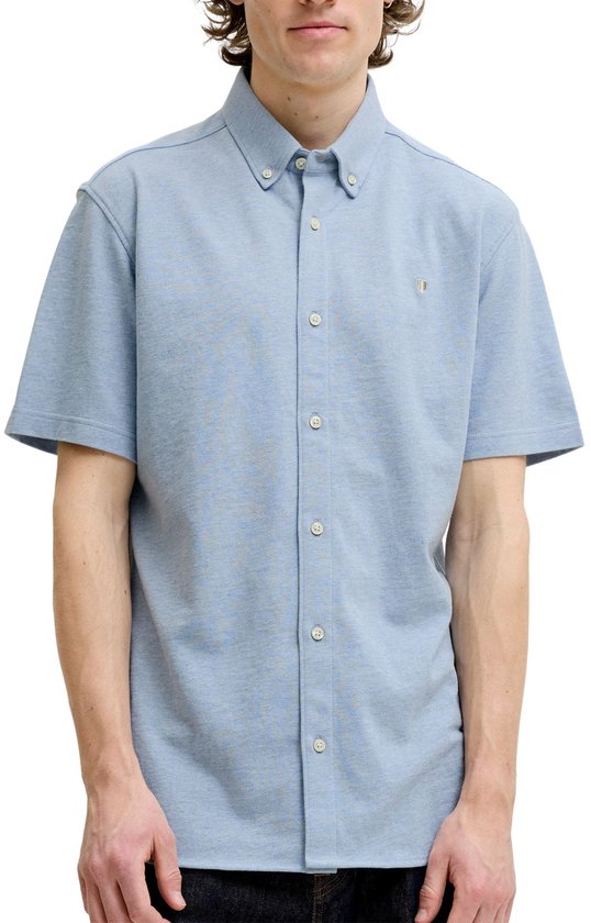 Man met krullend haar draagt een lichtblauwe korte mouw polo, staand op witte achtergrond.