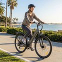 Vrouw in casual kleding, helm en zonnebril, rijdend op een grijze elektrische fiets langs de kust.