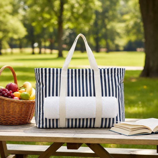 Blauw-wit gestreepte strandtas met opgerolde witte handdoek op picknicktafel naast fruitmand en boek.