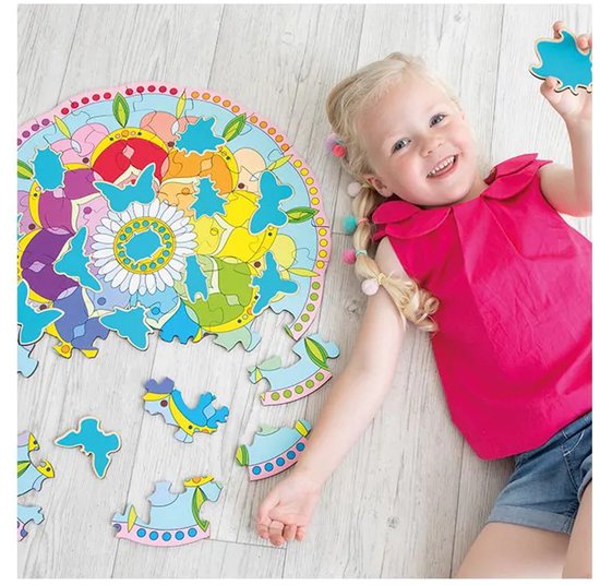 Une fille souriante aux couettes blondes avec des pompons est allongée sur le sol à côté d'un puzzle rond, avec des pièces détachées de chevaux et de bots. Le puzzle est un mandala avec des fleurs.