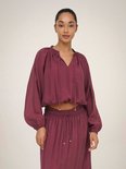 Model draagt een bordeauxrode crop top met pofmouwen en een v-hals met strikdetail. De top is gecombineerd met een bijpassende rok.